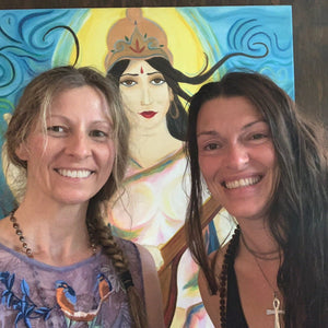 Two divine humans smiling in front of a painting of Saraswati. 